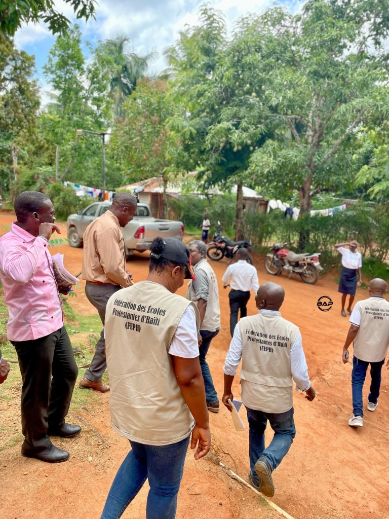 Sur une route en terre rouge : groupe en marche, gilets FEPH au dos siglés Fédération des Écoles Protestantes d'Haïti, végétation luxuriante