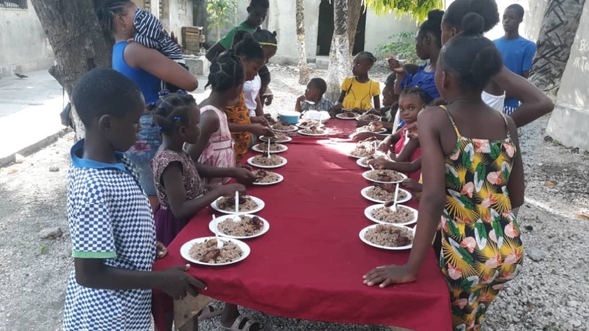Repas collectif en plein air : enfants et adultes autour d'une longue table, assiettes de riz et haricots