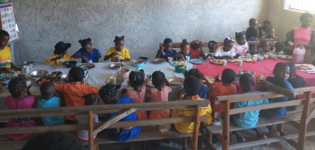 Élèves en uniforme prenant un repas à la cantine scolaire, équipes en tablier servant depuis de grandes marmites