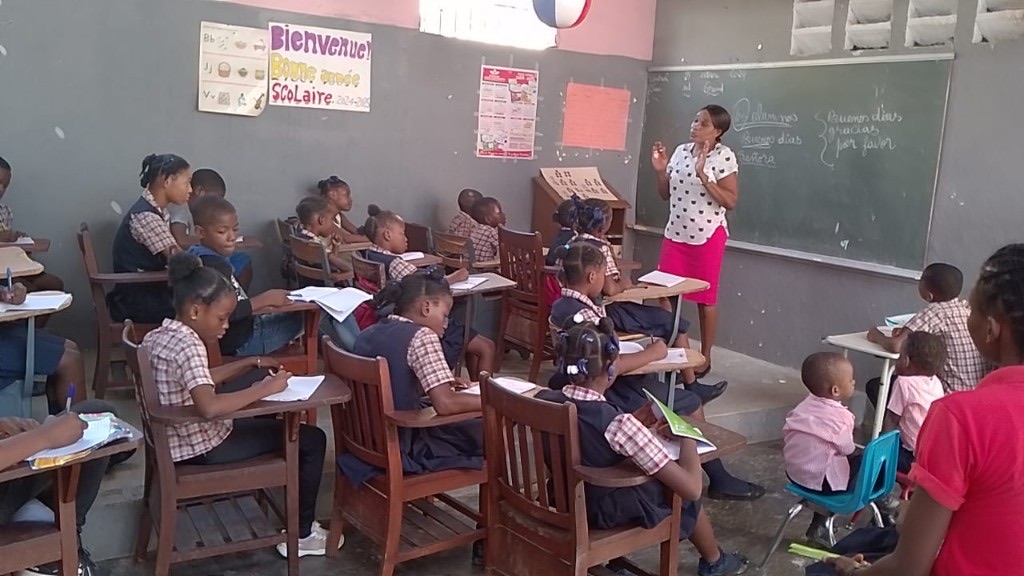 Salle de classe : enseignante au tableau, élèves en uniforme bleu, affiches pédagogiques et carte d'Haïti sur les murs